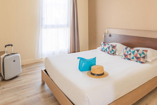 Chambre d'hôtel de la gamme classique avec un grand lit double doté de coussins colorés, une valise à roulettes blanche et un sac à main turquoise avec un chapeau de paille posé dessus, évoquant une atmosphère accueillante pour les voyageurs.