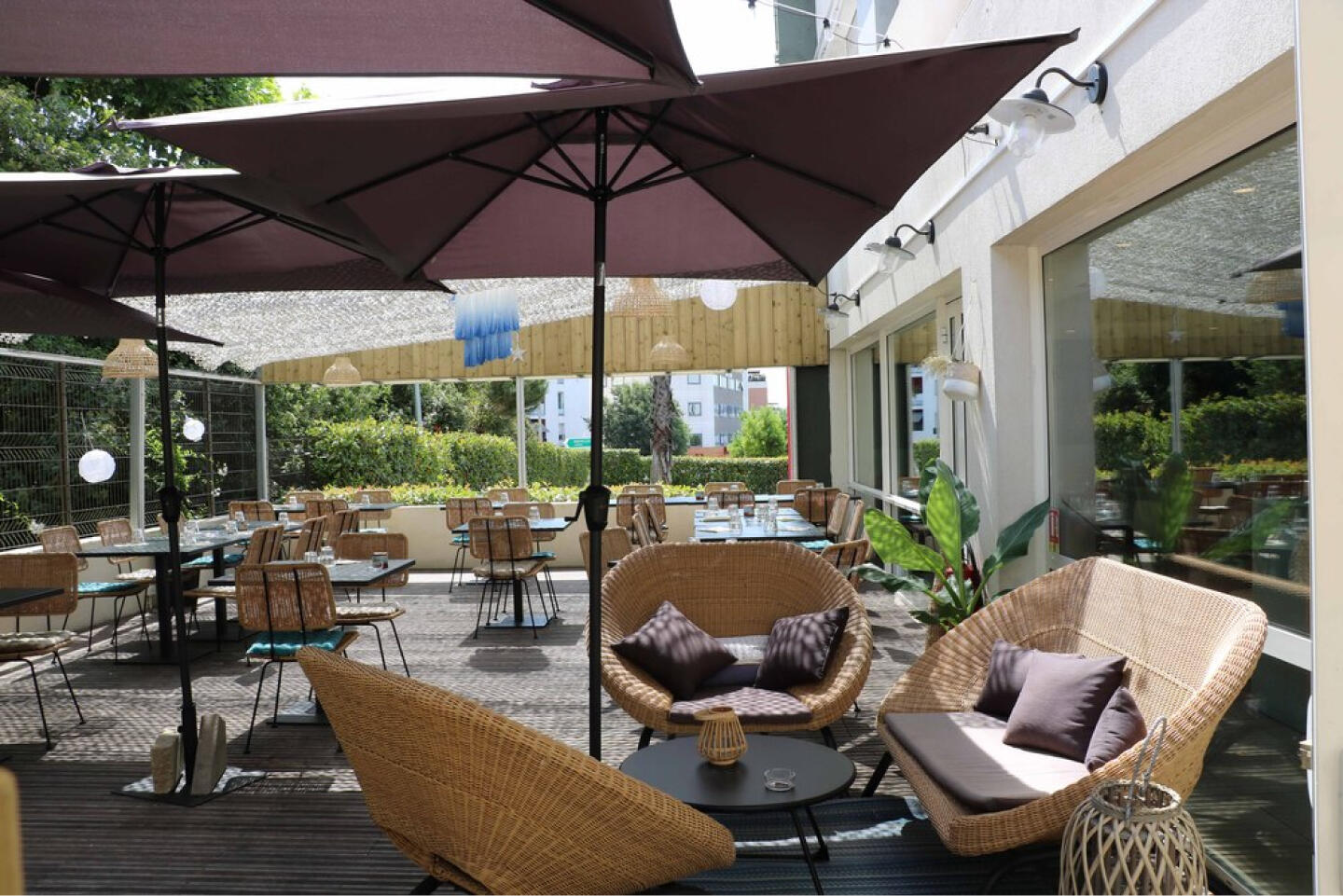 Terrasse extérieure du Restaurant Bistrot City Montpellier Ovalie avec des fauteuils en osier confortables sous un grand parasol marron, des tables dressées pour le repas et des plantes vertes, offrant un cadre détendu pour un repas en plein air.