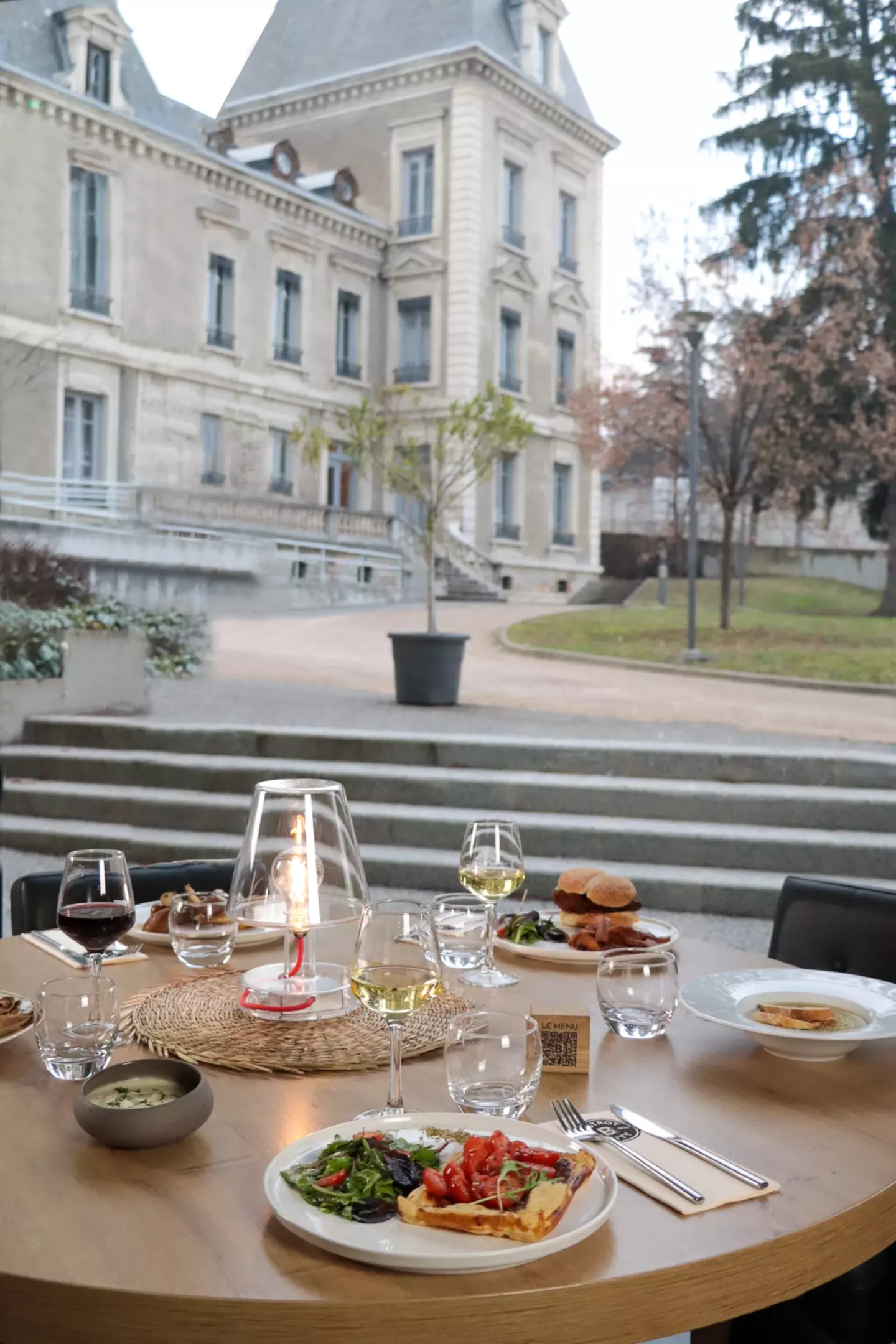 Vista de una mesa en la terraza del Bistrot City en la Cité Internationale de Lyon, preparada con platos de comida gourmet, copas de vino tinto y blanco, una linterna encendida, con un gran edificio clásico al fondo.