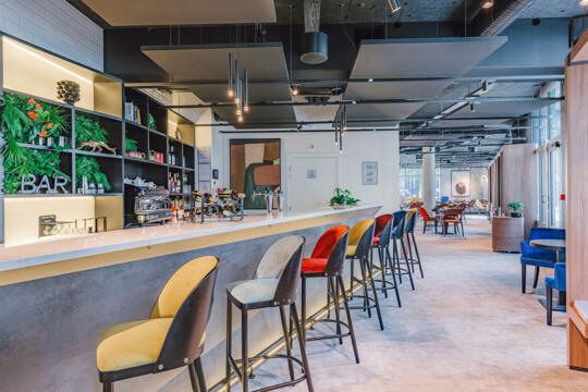 Modern hotel bar with concrete counter, high stools with multicolored velvet seats, black shelves with plants and bottles, professional coffee machine, industrial hanging lights, dining area in the background with wooden tables and comfortable chairs, urban and relaxed atmosphere