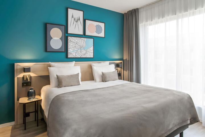 Contemporary hotel room with a large central double bed covered with a grey duvet, matching beige headboard, taupe pillows and decorative cushions, turquoise blue accent wall behind the bed adorned with modern artistic frames, two bedside wall lamps, sheer grey curtains and window allowing natural light in