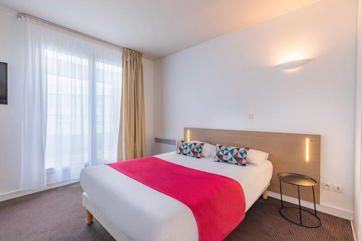 Modern and comfortable room with double bed, pink bed linen, decorative pillows, and soft lighting at an Appart’City aparthotel in Lyon.