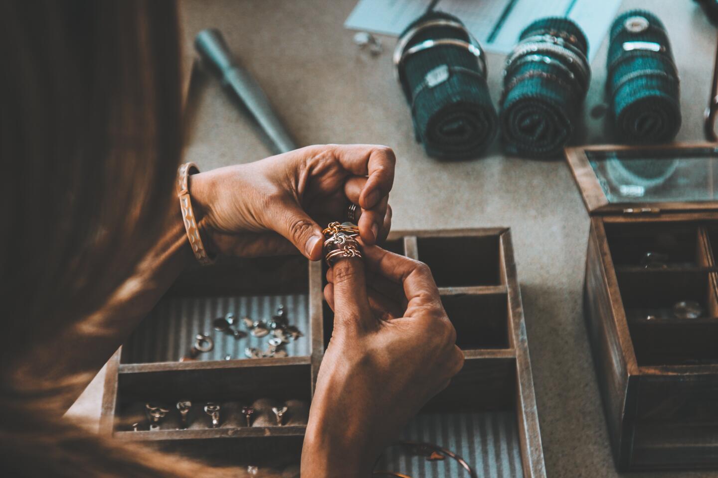 Jewelry artisan meticulously working on a ring in their workshop, with tools and compartmentalized boxes containing various pieces of jewelry in the background, conveying an ambiance of creativity and precision for the Bijorhca Paris exhibition.