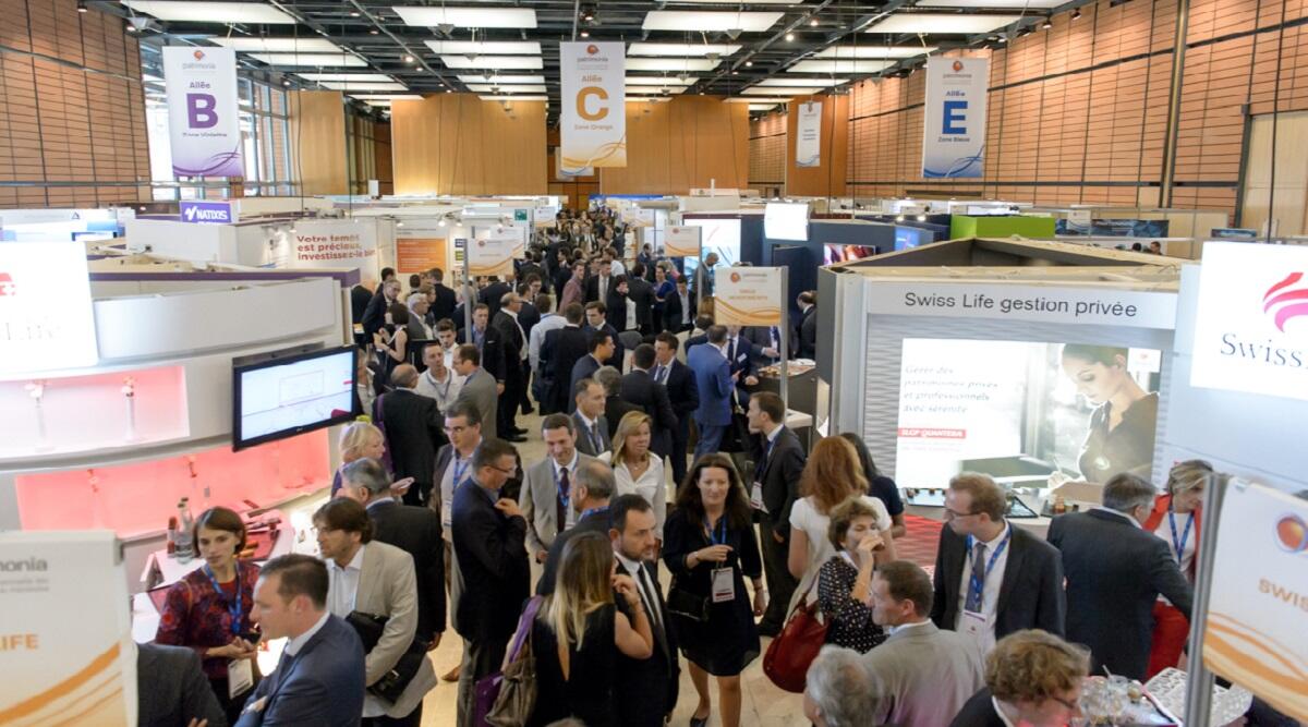 Aerial view of a bustling exhibition hall at the Patrimonia convention, with professionals in suits navigating between various company booths like Swiss Life. The aisles are crowded with visitors engaged in conversations and consultations.