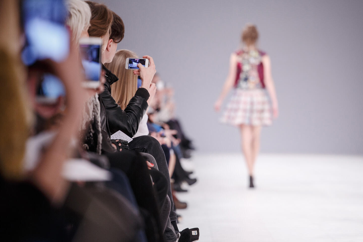 Audience members capturing a fashion show on their smartphones at Paris Fashion Week. In the foreground, a row of seated people, some snapping photos of a model in a short, colorful dress walking on the illuminated runway.