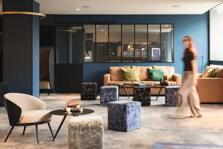 Blue lounge area with tan leather sofa, green cushions, cube ottomans and a woman walking in the background