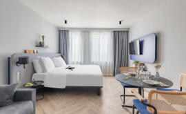 Vista interior de una suite Appart'City Collection Paris Roissy con cama queen-size, ropa de cama blanca, TV montada en la pared, área de comedor con mesa baja y sillas de mimbre, y un sofá gris en un decorado neutro y luminoso.