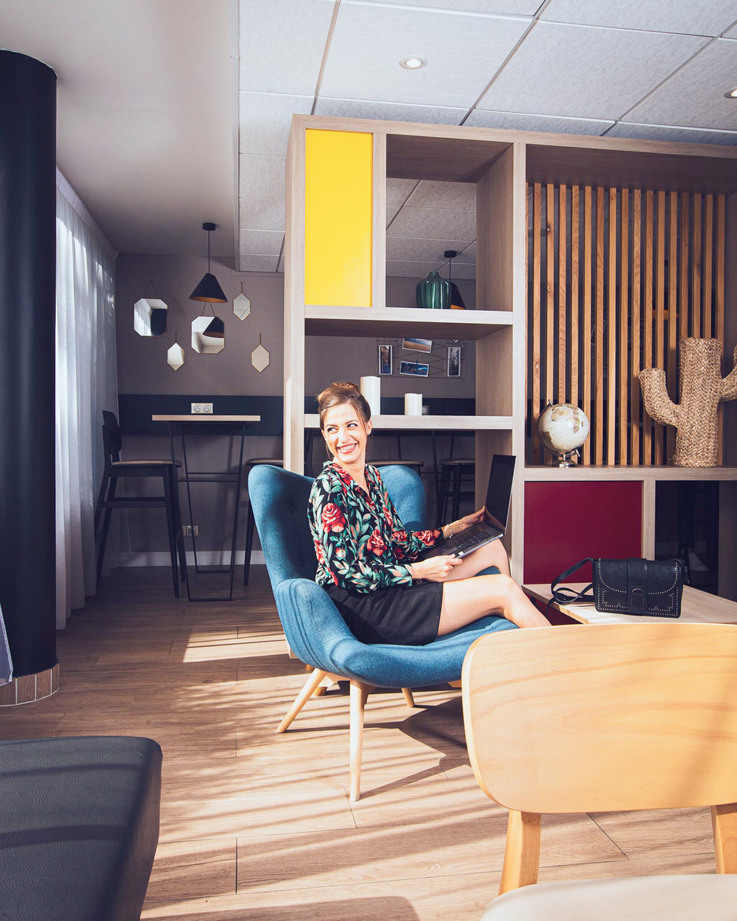 Smiling woman working on a laptop while sitting comfortably in a blue fabric armchair in an Appart'City apartment, with a modern interior that includes wooden shelving, a workspace desk, and stylish home decor.
