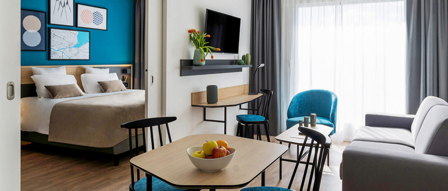 Zeitgenössisches Interieur eines aparthotel der  Collection Reihe bei Appart'City, mit einem Doppelbett, Wandkunst, einem kompakten Arbeitsplatz, einem bequemen Sofa, einem Couchtisch mit einer Obstschale und stilvoller Dekoration in Blau- und Grautönen.