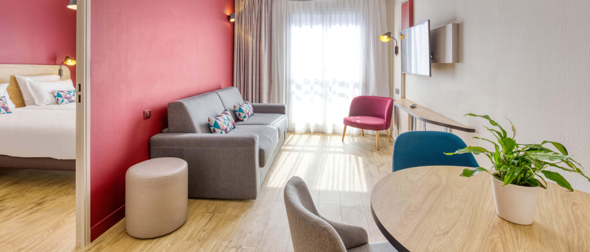 AC Confort apartment at Appart'City with a living room featuring pink walls, a gray sofa, and a beige pouf, a separate bedroom with a cozy bed, a desk with colorful chairs, modern decor, and a green plant adding a touch of nature.