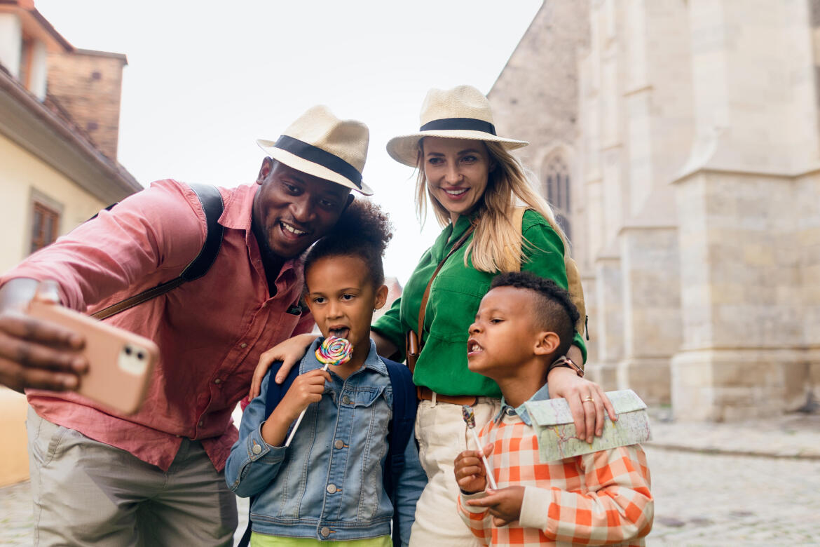 Glückliche Familie im Urlaub beim Selfie machen, mit einem lächelnden Vater, Mutter und zwei Kindern mit Hüten, die vor historischen Gebäuden stehen, was den Spaß an Familienaufenthalten verdeutlicht, die von Appart'City angeboten werden.