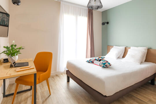 Chambre d'appart'hôtel Appart'City lumineuse et contemporaine avec un grand lit confortable, une chaise de bureau orange vive, un espace de travail accueillant, et des touches de couleur pour un séjour relaxant à Lyon.