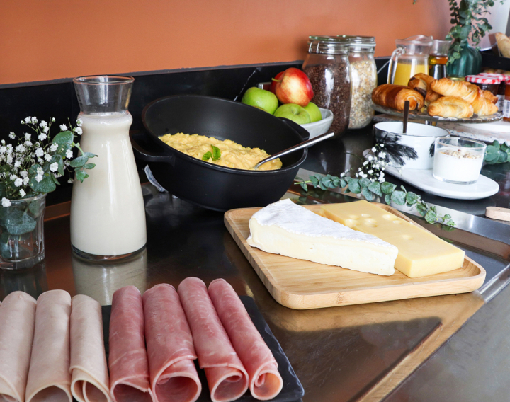 Buffet de desayuno en un apartahotel AC Classic con rodajas de jamón, queso en una tabla de madera, huevos revueltos, una jarra de leche, frutas frescas, cereales y una selección de bollería en una encimera negra.