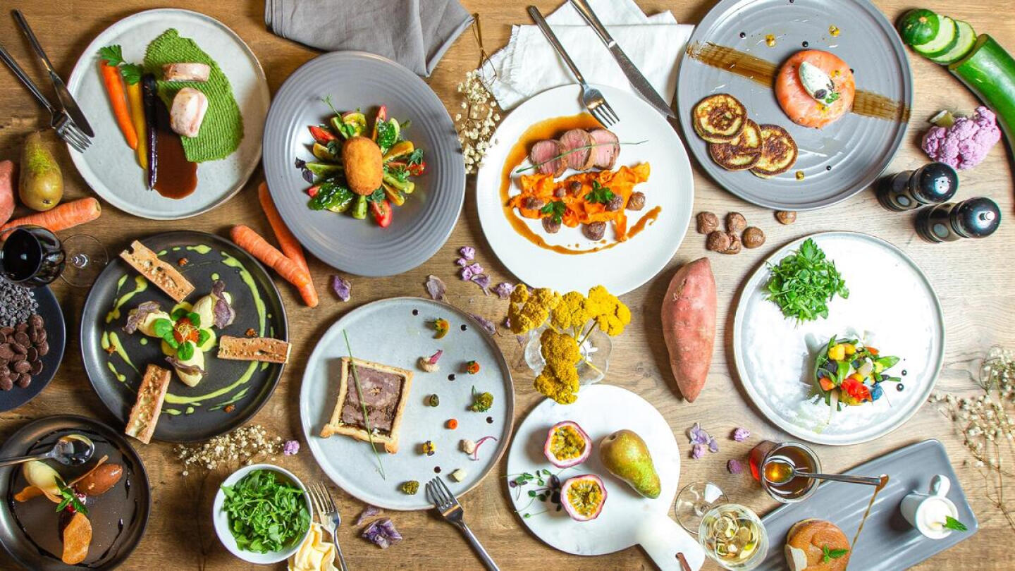 Array of colorful, artistically presented gourmet dishes on a wooden table at Bistrot City, featuring a variety of vegetables, meats, and seafood, complemented with sauces, fresh herbs, and spices, alongside cutlery and two glasses of wine.
