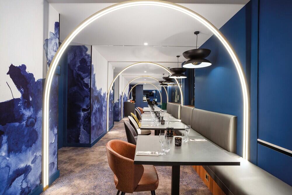 Interior moderno de Bistrot City Aeropuerto de Ginebra Ferney-Voltaire con una fila de sillas de terciopelo y mesas preparadas, iluminado por arcos luminosos y elegantes lámparas colgantes, con paredes adornadas con arte abstracto azul.