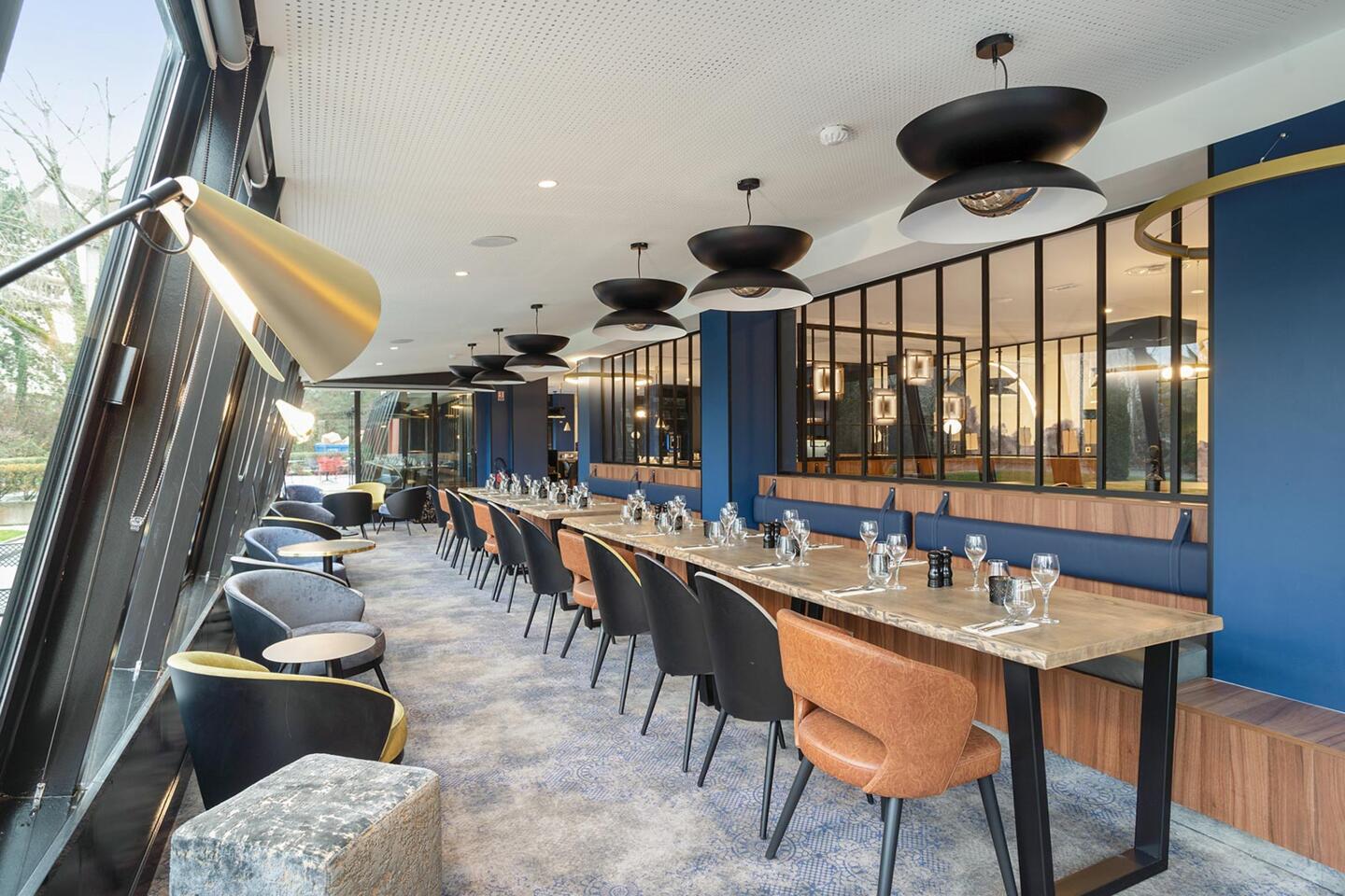 Chic dining space at Bistrot City Geneva Airport Ferney-Voltaire with gray velvet seating and brown leather stools, set wooden tables, designer suspended lamps, and a large window overlooking the outdoors.