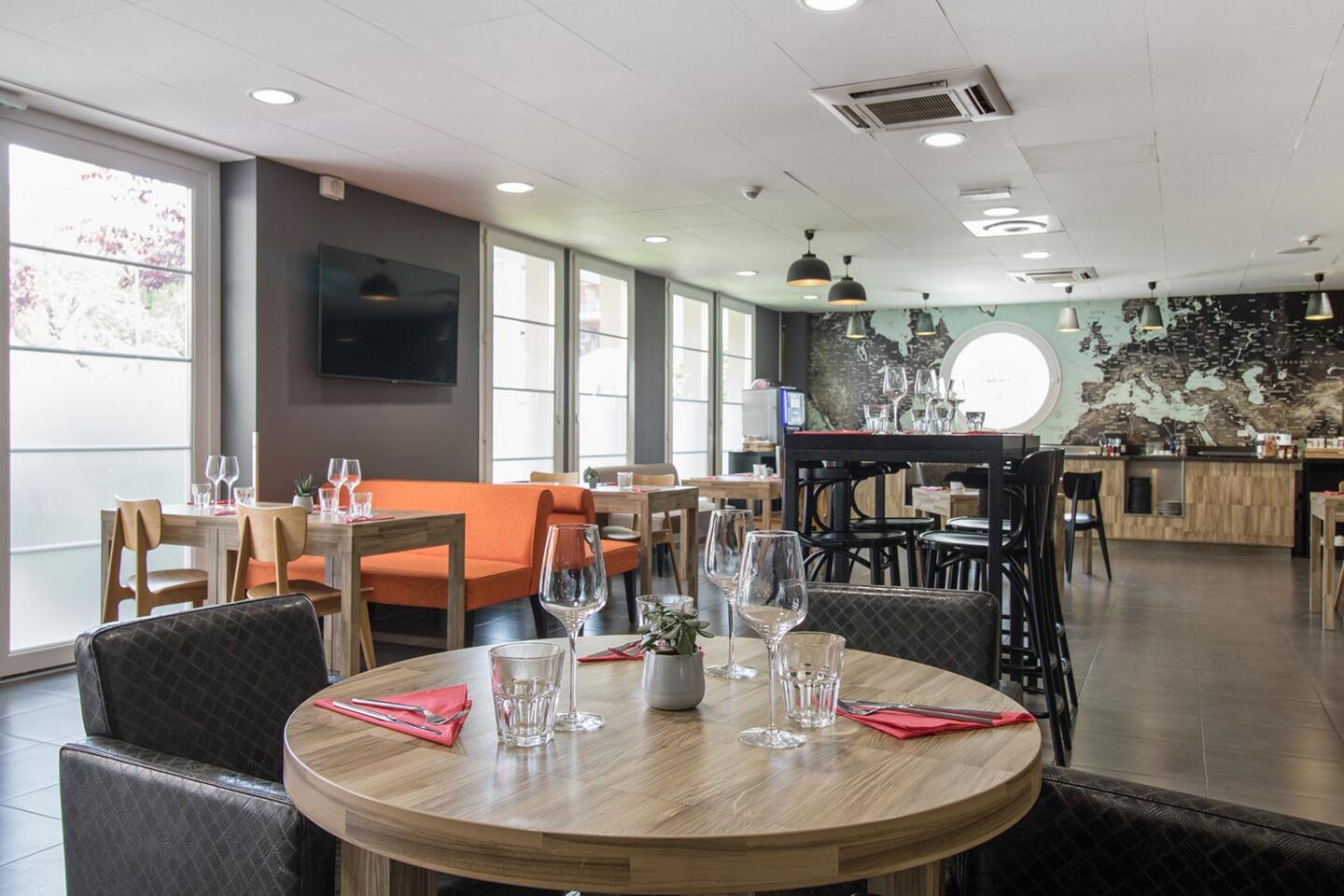Bright and modern interior of Bistrot City Le Bourget featuring round and square set tables, glasses and red napkins, a wall adorned with a world map, under stylish pendant lighting, and beside large windows.