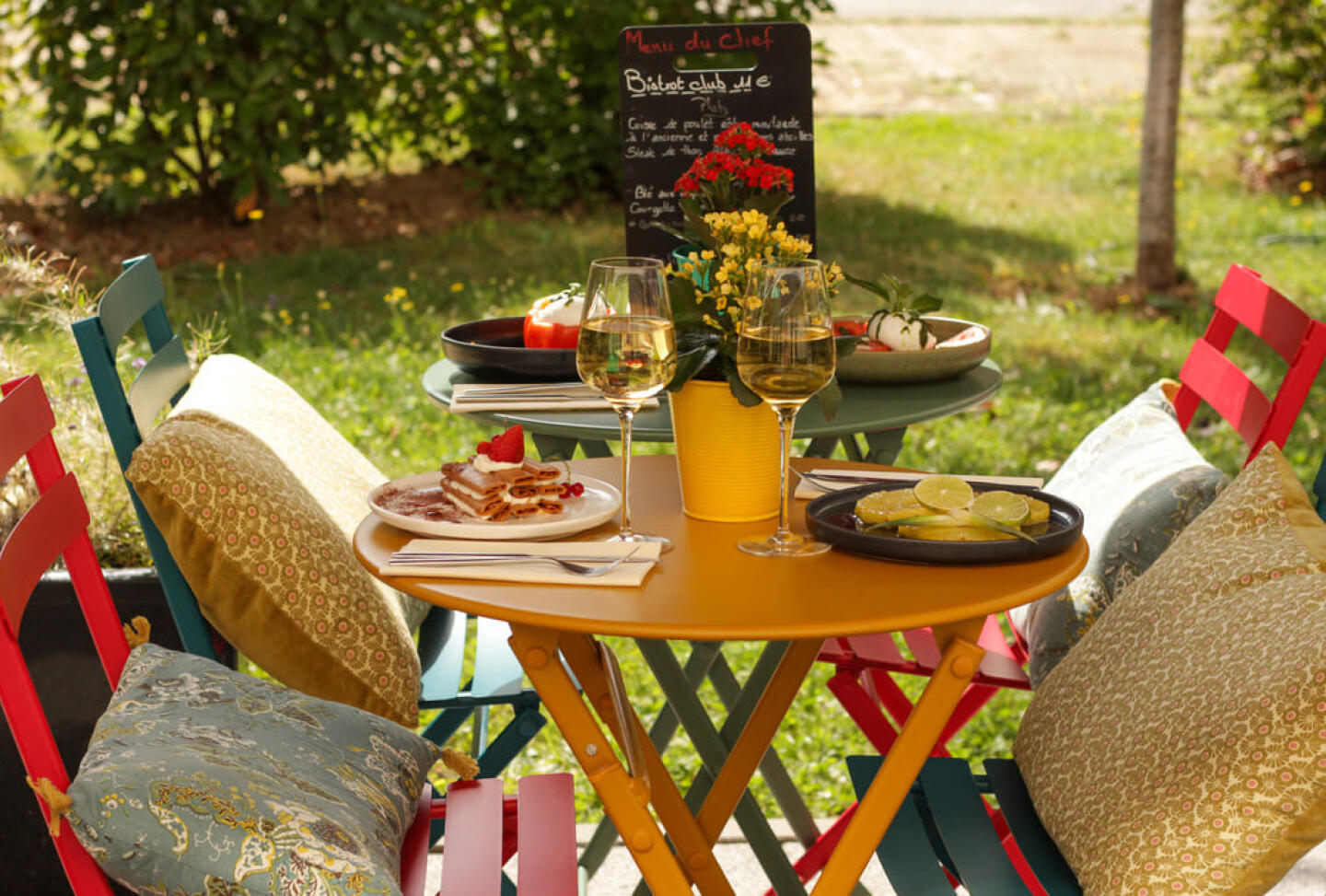 Table de bistrot colorée avec des verres de vin blanc, une assiette de crêpes garnies et un bol de fruits frais, sous un tableau noir affichant le menu, entourée de verdure dans l'ambiance conviviale du Bistrot City Le Bourget