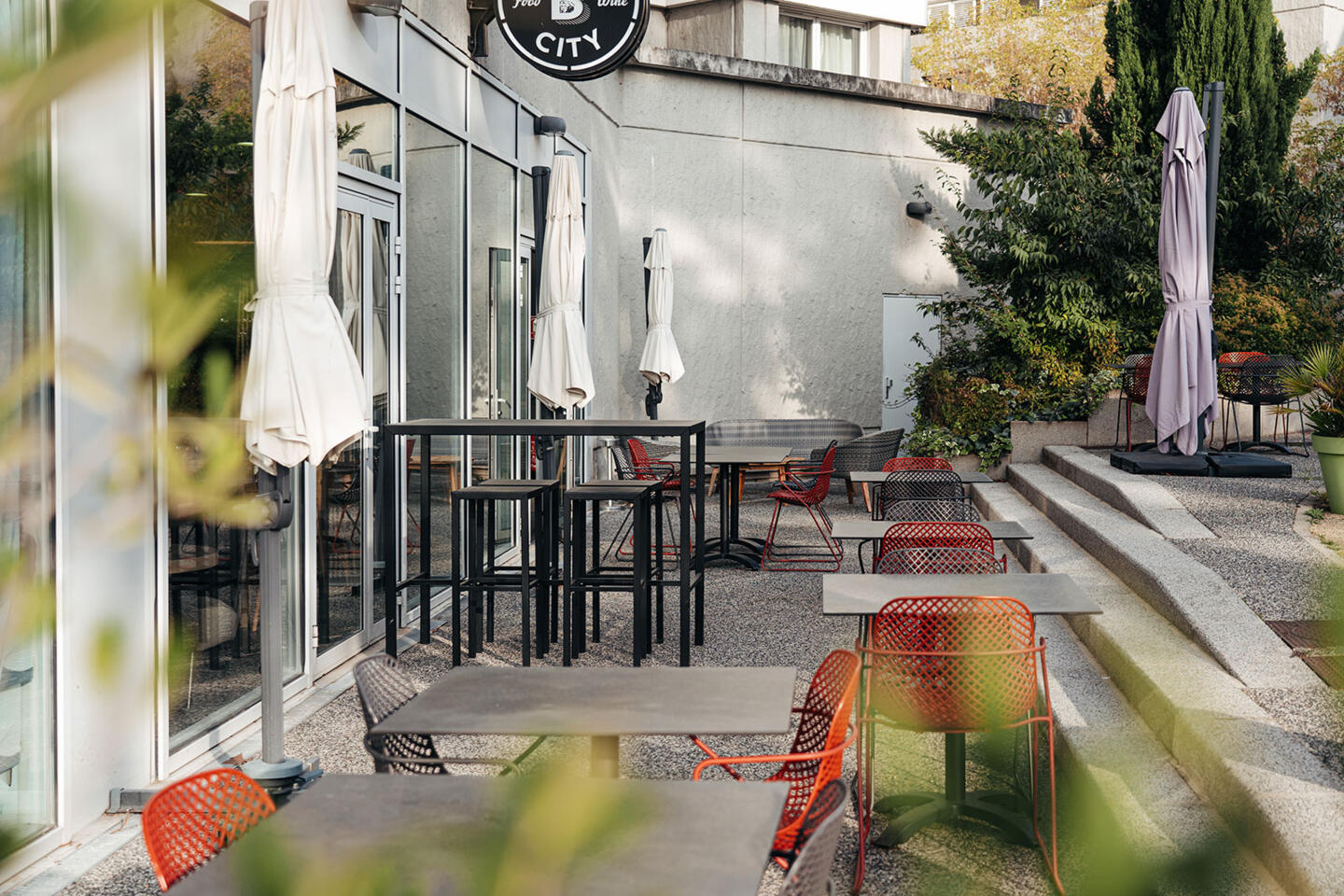 Ruhige Terrasse des Restaurants Bistrot City in Lyon Cité Internationale mit modernen schwarzen Metalltischen und Stühlen mit orangefarbenen Sitzen, geschlossenen Sonnenschirmen, in einem städtischen Umfeld mit grüner Bepflanzung, das zur Entspannung einlädt.