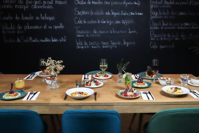 Table de restaurant élégante avec des plats gastronomiques variés, verres de vin rouge et blanc, décorée de fleurs sauvages, avec un tableau noir affichant le menu du jour en arrière-plan.