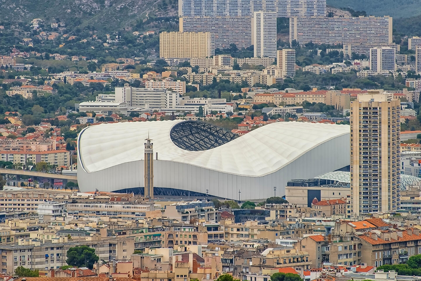 Aerial view of the Stade Vélodrome in Marseille, with its iconic white wave-shaped structure, surrounded by the city's dense urban architecture and hills in the background, suggesting an immersive experience for visitors staying in local aparthotels.