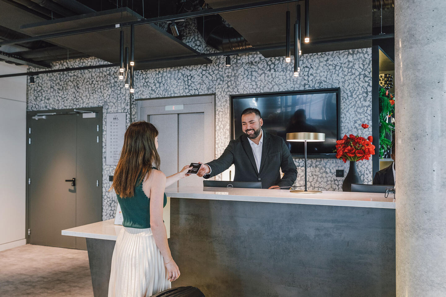 Male receptionist in a suit handing over a room card to a long-haired female guest in a bright and stylish Appart'City lobby, highlighting the hospitality and comfort of aparthotels.