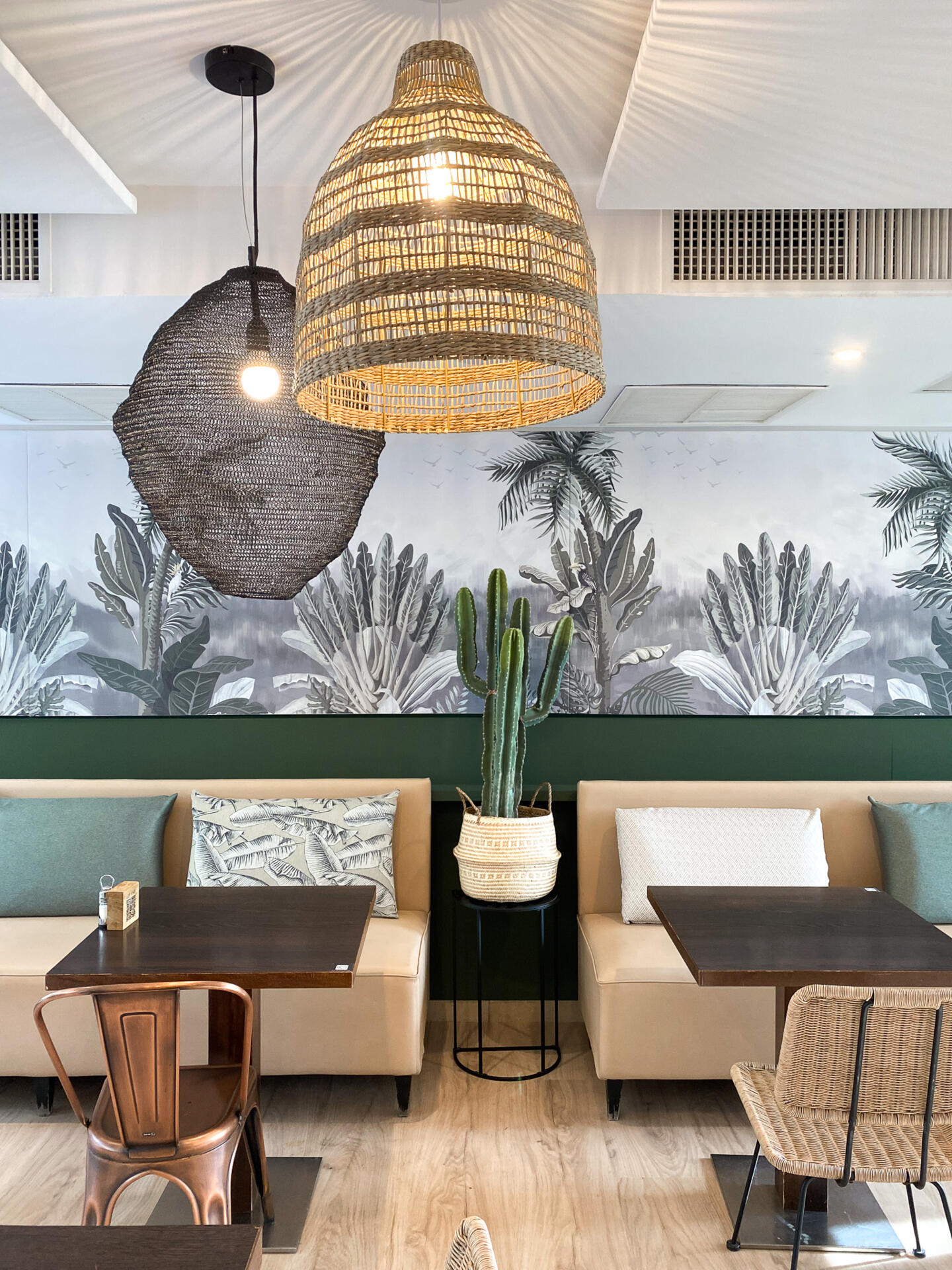 Interior decor of Bistrot City Montpellier Ovalie, featuring a rattan pendant light, a wall adorned with a tropical cactus mural in shades of grey, and a welcoming seating area with benches and printed cushions.