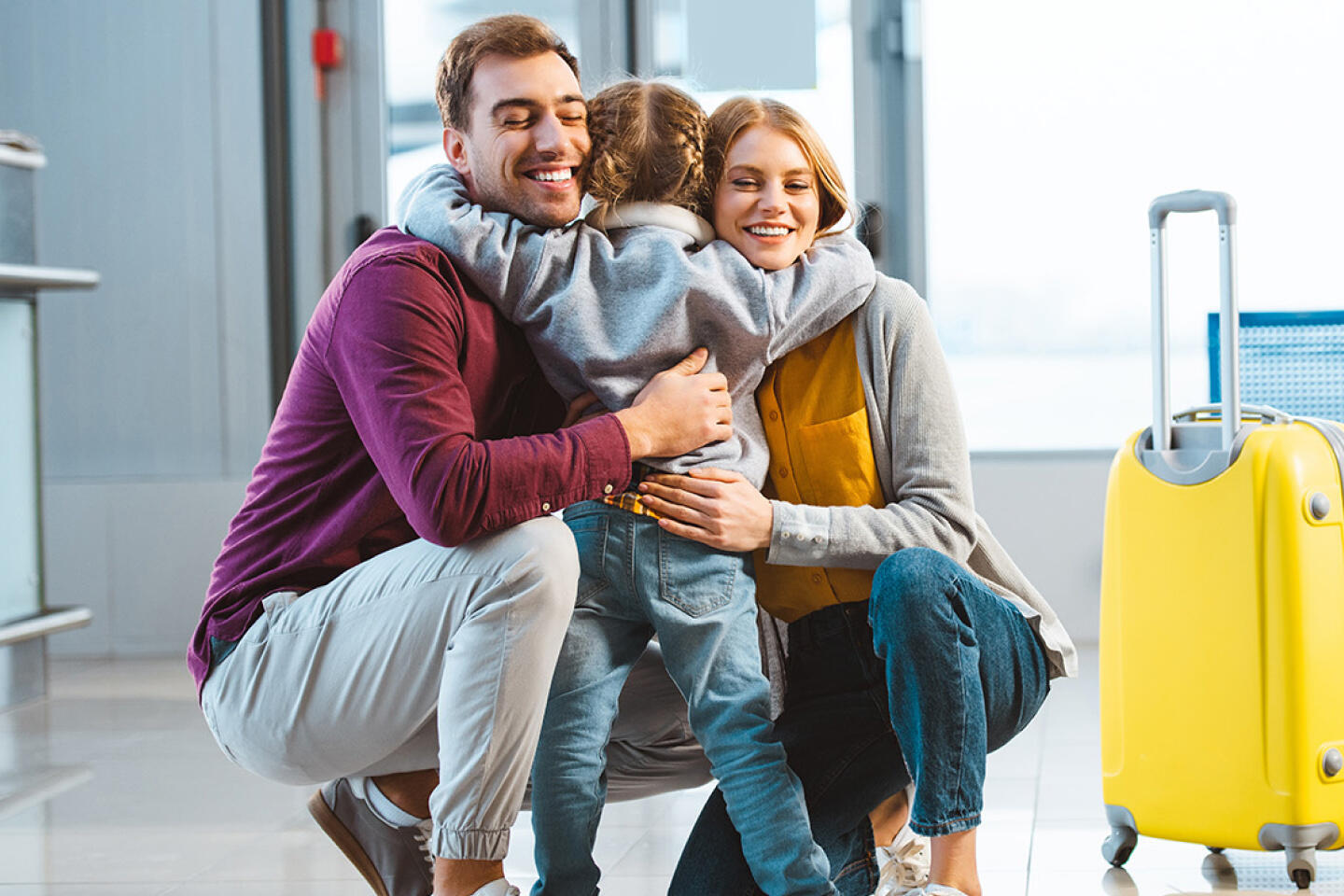 Glückliche Familie mit einem Kind, das sich freudig im Flughafen Marseille Vitrolles umarmt, neben einem leuchtend gelben Koffer, der sorgenfreies Reisen symbolisiert.