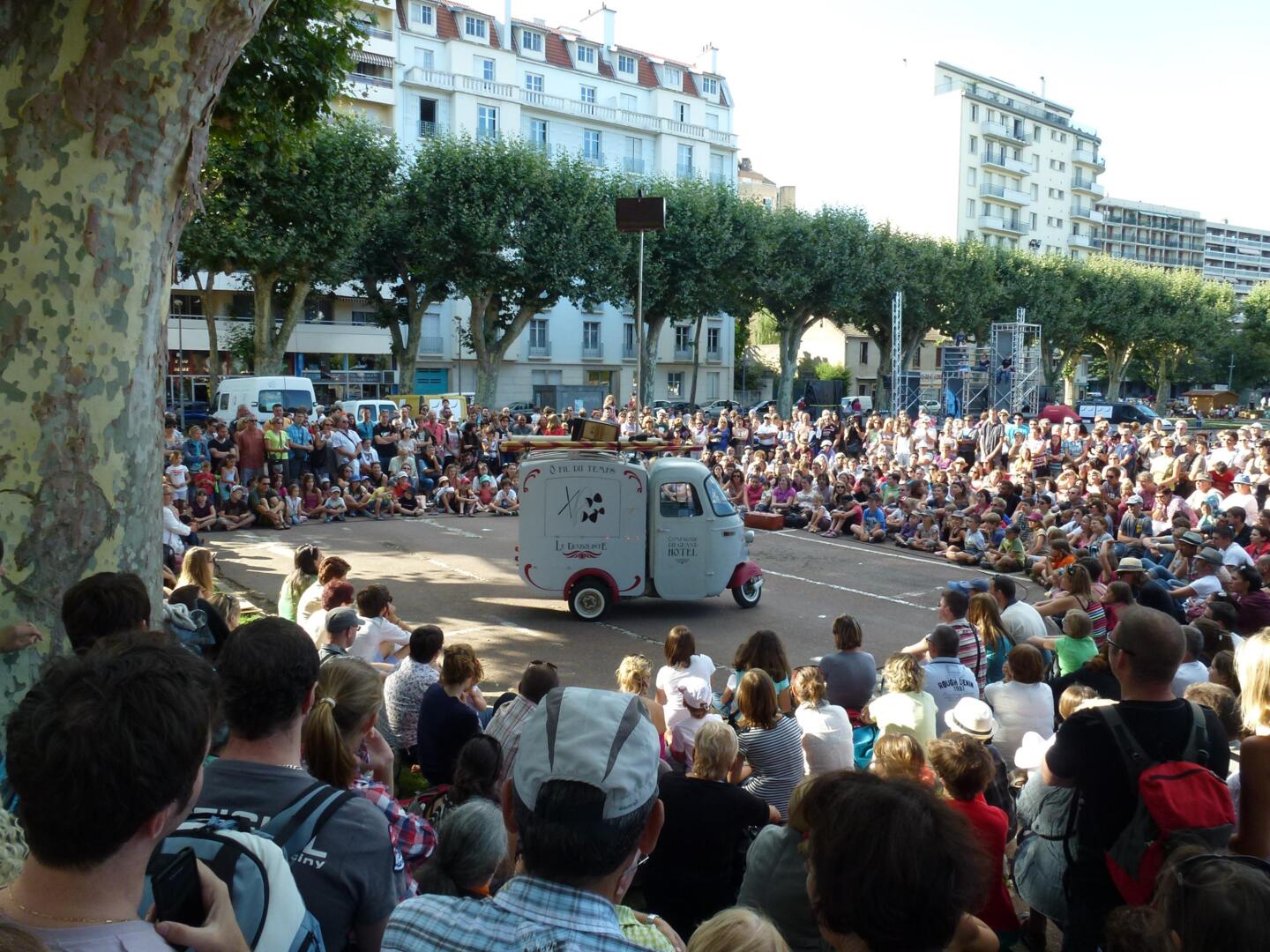 Público cautivado asistiendo a una actuación callejera en el festival 'Chalon dans la Rue', con un vehículo de espectáculo estacionado frente a los espectadores sentados.