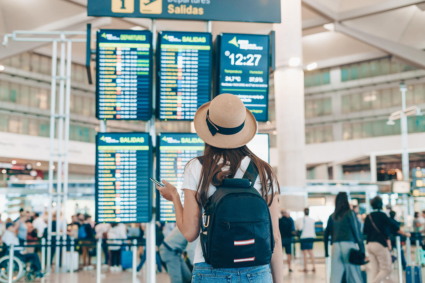 Mujer de espaldas con sombrero y mochila consultando su teléfono frente al tablón de salidas del aeropuerto de Burdeos.