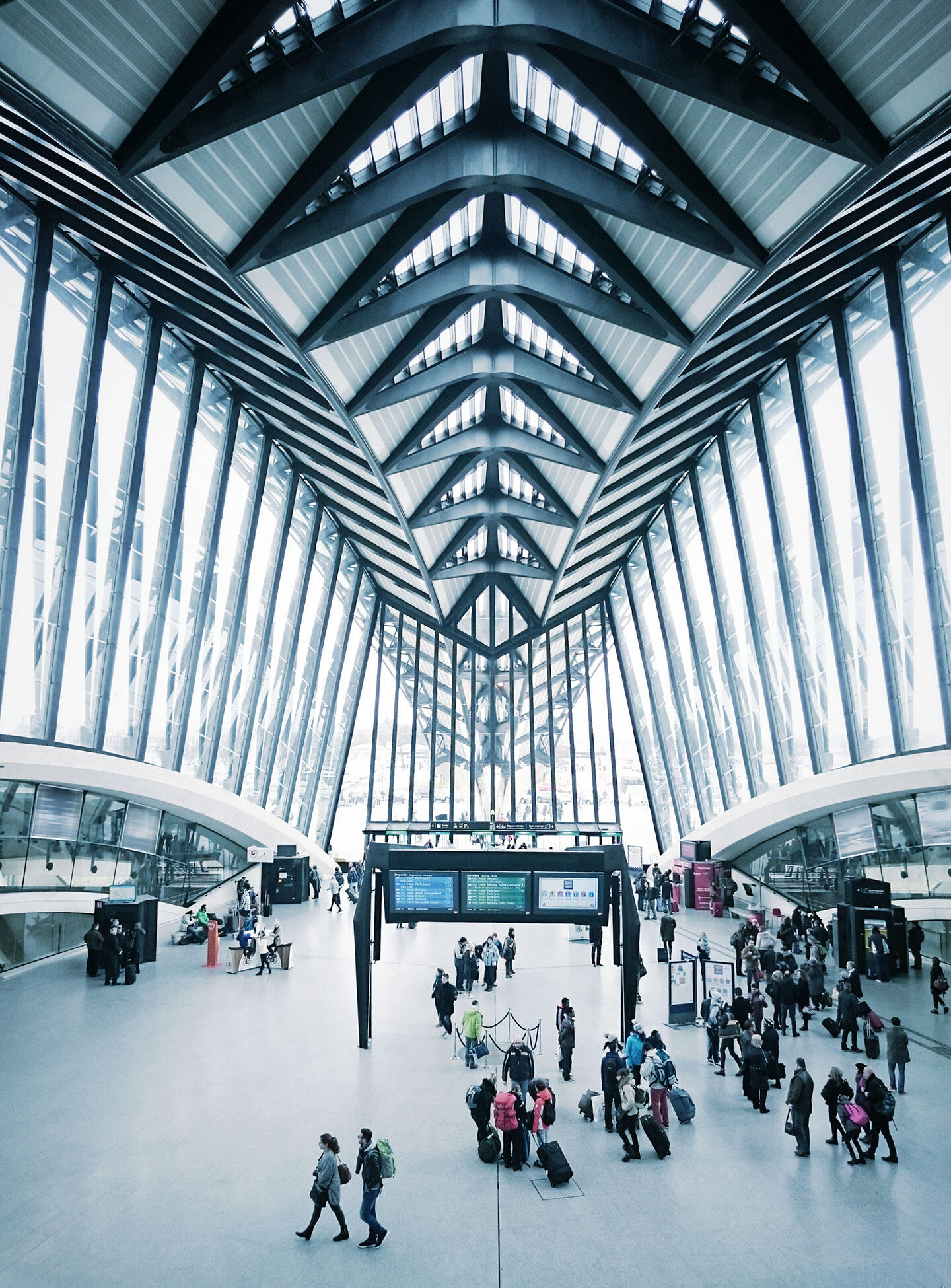 Innenansicht des Terminals am Flughafen Lyon mit Reisenden und Informationsbildschirmen unter einer modernen gewölbten Glasdecke.