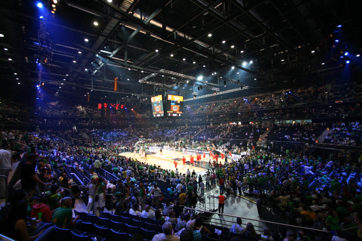 Vista interior de un pabellón deportivo abarrotado durante un partido de baloncesto, con los espectadores vestidos de verde y la acción en el centro, en el suelo iluminado.