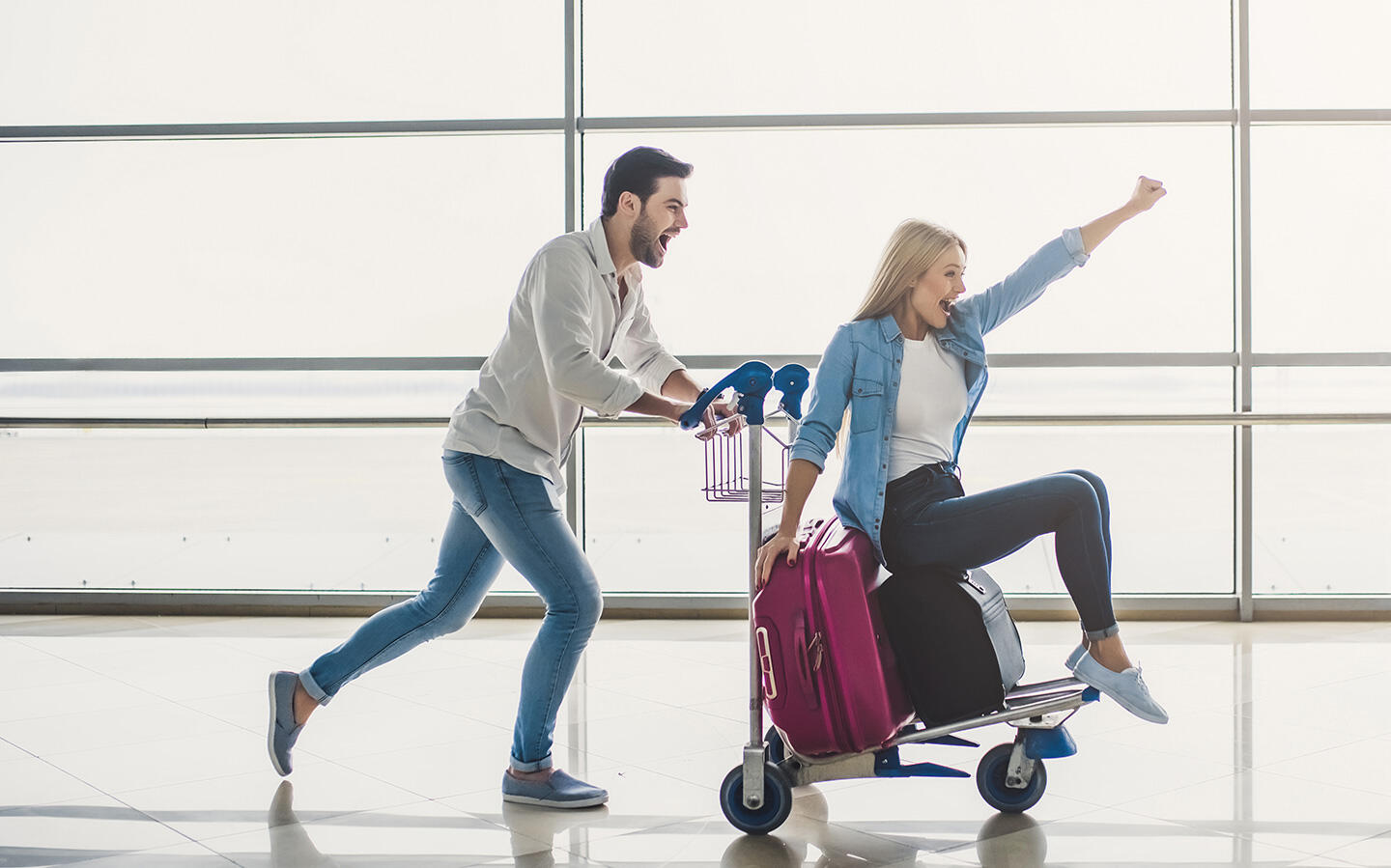 Fröhliches Paar, das sich mit seinem Gepäck am Flughafen Nantes beeilt, Mann, der eine lächelnde Frau auf einem Gepäckwagen schiebt.