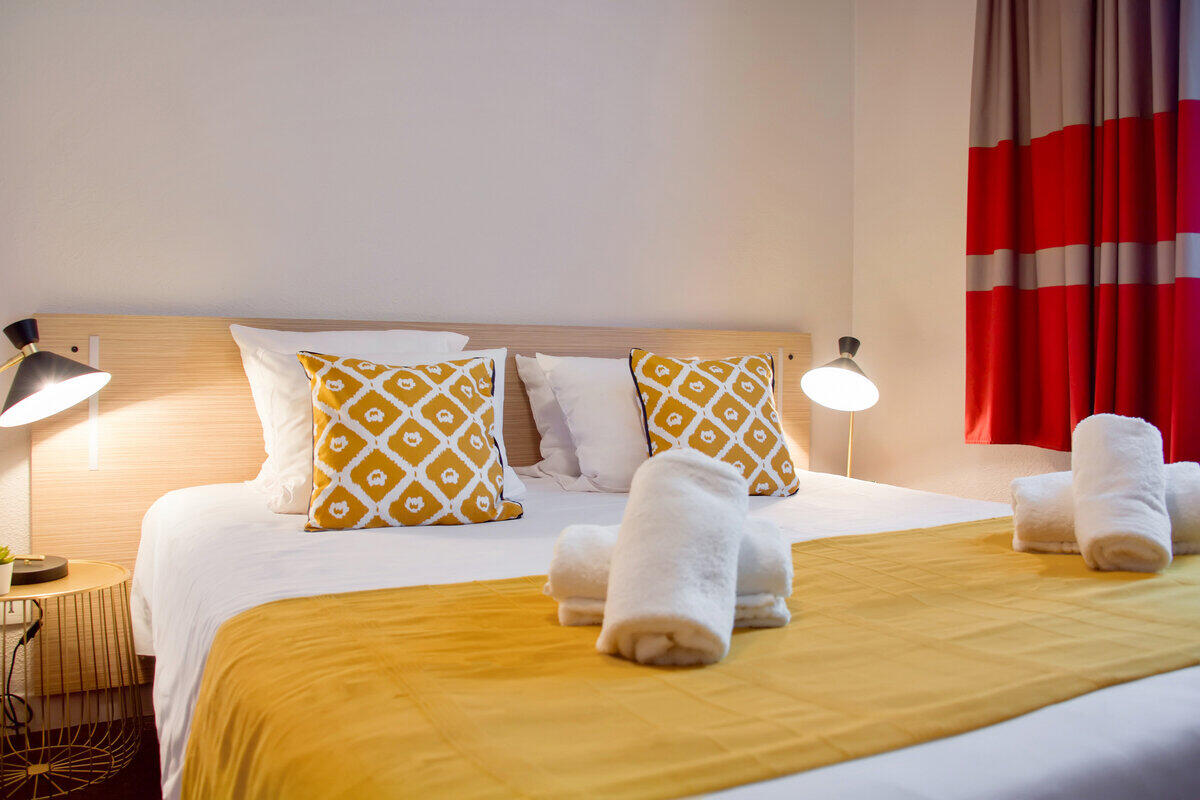 Moderna y acogedora habitación de apartamento Appart'City con cama de matrimonio, cojines decorativos amarillos, sábanas blancas y toallas de rizo enrolladas.