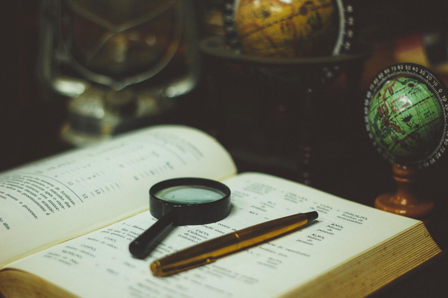 Antique book open with magnifying glass and pens, globes in the background, evoking historical exploration for the Rendez-Vous de l'Histoire.