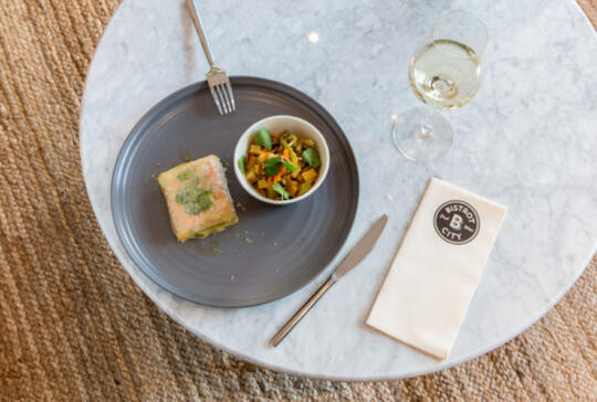 Overhead view of a terrine dish and a vegetable salad in black bowls on a marble table with a Bistrot City logo napkin, accompanied by a stainless steel fork and knife and a glass of white wine.