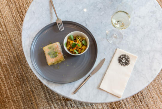 Vue en plongée d'un plat de terrine et d'une salade de légumes dans des bols noirs sur une table de marbre avec une serviette et un logo Bistrot City, accompagnés d'une fourchette et d'un couteau en acier et d'un verre de vin blanc.