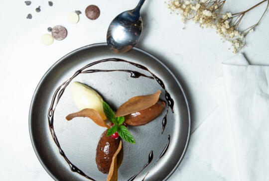 Top view of an artistic chocolate dessert on a grey plate, featuring a vanilla ice cream quenelle, chocolate leaf decoration, a chocolate sauce spiral, and a small green leaf, accompanied by a vintage spoon and dried petals.