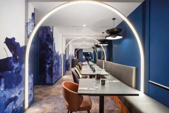 View of the corridor of Bistrot City Geneva Ferney-Voltaire with grey benches and brown velvet seats under the illumination of lighted arches, blue walls with abstract splashes, and black hanging lights.