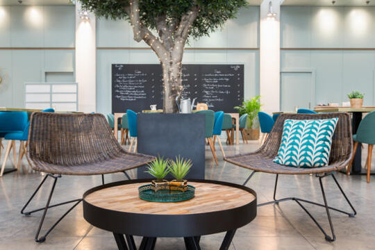 Espace détente du Restaurant Bistrot City Lyon Part Dieu avec des fauteuils en rotin, des coussins à motifs, une table basse ronde, un grand arbre intérieur, et un tableau noir avec le menu en arrière-plan éclairé par une lumière naturelle abondante.