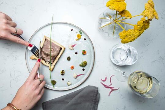 Draufsicht auf einen eleganten Tisch im Bistrot City Genf Flughafen Ferney-Voltaire mit einer weiblichen Hand, die eine Pastete umgeben von verschiedenen kleinen Beilagen auf einem modernen Teller schneidet, neben einem Glas Wasser und einem Cocktail auf einer Marmorunterlage.