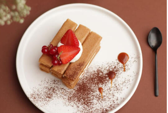 Elegant plate featuring rolled crepes topped with strawberries and red currants, dusted with cocoa, accompanied by a black spoon on a brown background, reflecting the fine patisserie offered by Bistrot City Le Bourget.