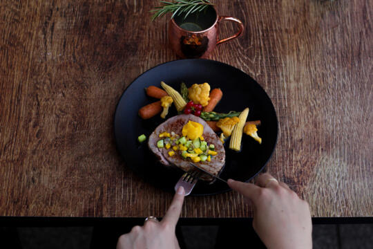 Refined main dish served at Bistrot City Le Bourget featuring sliced meat topped with colorful vegetables such as baby corn, asparagus, and carrots, with a touch of sauce, on a rustic wooden table with a hand using a fork.