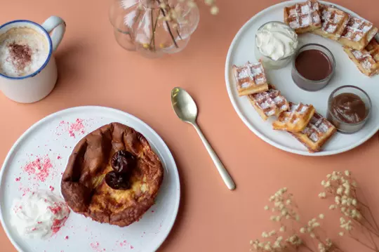 A cozy breakfast setup featuring a cappuccino with cocoa powder, a chocolate Dutch baby pancake with a cherry on top, waffles with powdered sugar alongside chocolate and caramel dips, and a decorative glass vase with dried flowers on a coral-colored table.