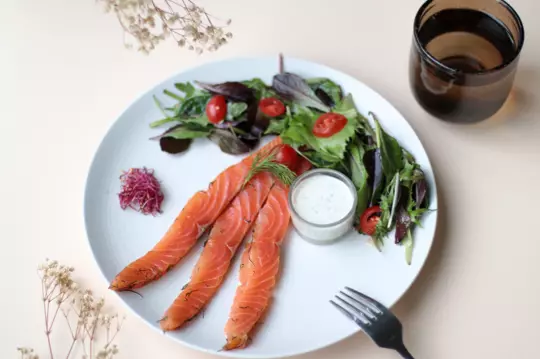 Plate of fresh salad and smoked salmon, accompanied by a dill sauce and petite vegetables, elegantly presented on a table at Bistrot City Lyon Cité Internationale, with a dark water glass and subtle floral decoration in the background.