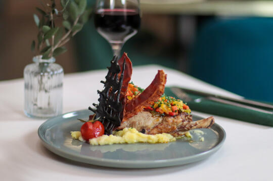 Plato refinado con chuleta de cordero adornada con brunoise de vegetales, sobre puré de papas, acompañado de una copa de vino tinto y decoración de crujiente pan negro.