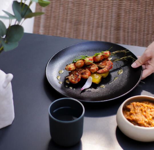 Spicy shrimp skewers on a black plate with sauce and lemon slices, presented on an elegant table.