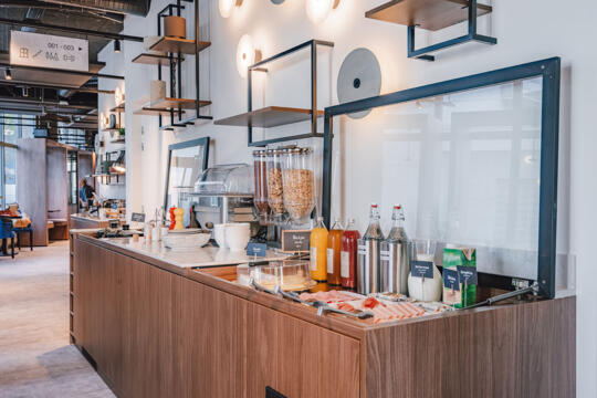 Área de buffet en un hotel con mostrador de madera marrón, surtido de jugos frescos en dispensadores transparentes, cereales en frascos, máquina de café profesional, bandejas de pasteles y frutas frescas, decoración elegante con estantes colgantes y luminarias modernas, ambiente amistoso y acogedor