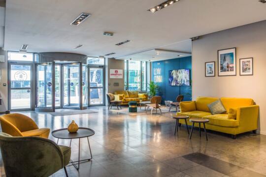 Lobby amplio y acogedor de Appart'City con cómodos sofás amarillos, mesas de centro elegantes y decoración mural artística, una invitación a relajarse después de un día en el Salon du 2 Roues en Lyon.