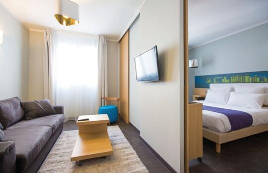 Appartement T2 lumineux et fonctionnel dans un appart'hôtel, avec un salon séparé équipé d'un canapé gris et une chambre accueillante pourvue d'un lit double, offrant un cadre de vie contemporain et confortable.